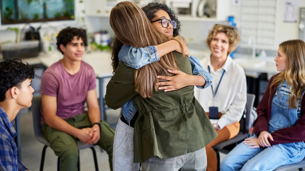 students hugging in class with teacher and colleagues in circle around them